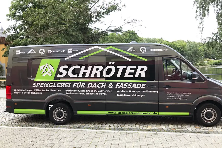 Dunkelgrauer Lieferwagen mit großer Beschriftung 'Schröter Spenglerei für Dach & Fassade' und weiteren Leistungsangeboten auf Kopfsteinpflaster an einem parkähnlichen Gewässer.