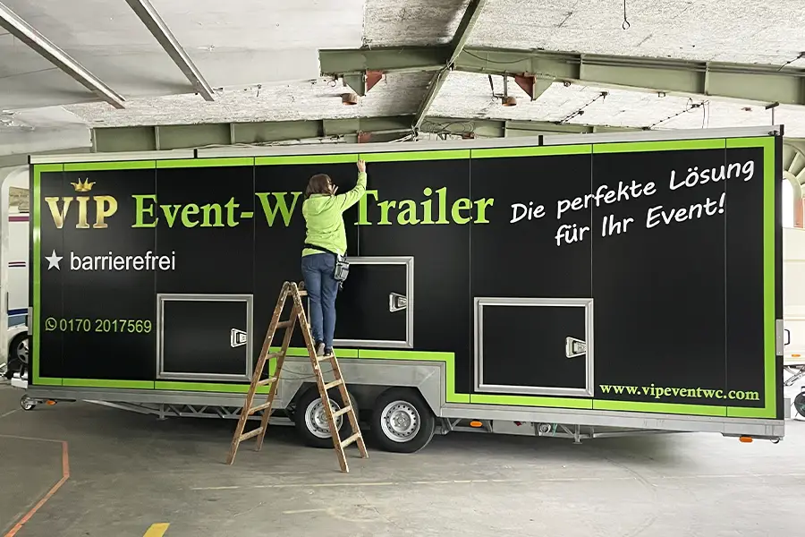 Frau auf einer Holzleiter beklebt einen schwarzen VIP Event-WC-Trailer mit grüner Schrift in einer Halle.