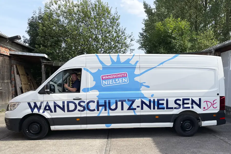 Weißer Lieferwagen mit blauem Farbklecks und Schriftzug 'Wandschutz Nielsen' sowie Fahrer im Führerhaus.