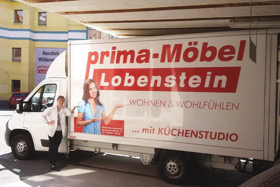 Frau steht neben weißem Transporter mit Werbung für prima-Möbel Lobenstein und Küchestudio.