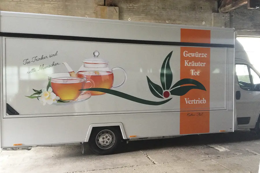 Weiße Lieferwagen-Seitenfläche mit Teeverkauf-Werbung, zeigt eine Glasteekanne, eine Teetasse und grüne Teeblätter mit dem Text 'Tee-Trinker sind nette Menschen' und 'Gewürze Kräuter Tee Vertrieb'.