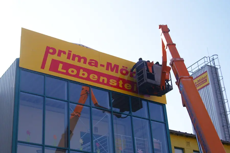 Zwei Arbeiter auf einer orangefarbenen Hebebühne befestigen ein gelbes Schild mit rotem Text an einem Gebäude mit Glasfassade.